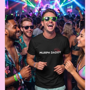 May include: A black short-sleeved crew neck t-shirt featuring the white text MURPH DADDY printed across the chest, accompanied by a small red graphic of lips positioned above the word DADDY. The shirt is made of a solid black fabric.