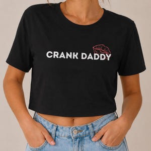 Może przedstawiać: Czarny krótki top z krótkim rękawem z białym napisem CRANK DADDY na piersi, któremu towarzyszy mały czerwony kontur ust umieszczony nad literą Y w słowie DADDY, zestawiony z jasnoniebieskimi dżinsami.