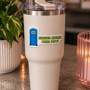 Può includere: Un bicchiere bianco in acciaio inossidabile con coperchio trasparente e cannuccia. Un adesivo sul davanti mostra un bagno portatile blu con il testo 2026 sopra la porta e il testo verde SURVIVED ANOTHER PORTA POTTY a destra del bagno.