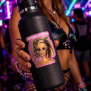 May include: A matte black insulated water bottle featuring a rectangular sticker with a pink border. The sticker displays a doll head with messy blonde hair and the text UNSTABLE BUT FUN printed in white capital letters on a pink background.