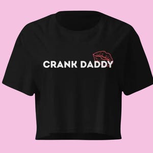 May include: Black crop top with the text "CRANK DADDY" in white, and a red lip graphic. The shirt has a crew neck and short sleeves. The background is a light pink color.