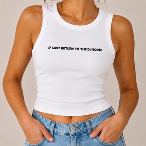 May include: A white ribbed tank top featuring the black text IF LOST RETURN TO THE DJ BOOTH printed across the chest, paired with light blue denim jeans. The sleeveless top has a round neckline and a fitted silhouette that ends at the waist.