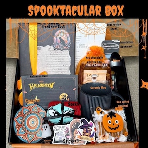 May include: A Halloween-themed gift box filled with a book, mug, socks, stickers, hot chocolate, and other treats. The box includes a Halloween card and a coaster. The text "SPOOKTACULAR BOX" is at the top.