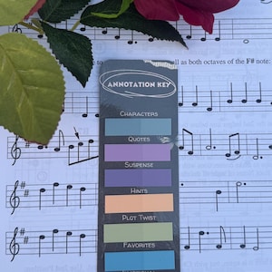 May include: A bookmark with the text "ANNOTATION KEY" and colorful labels for "CHARACTERS", "QUOTES", "SUSPENSE", "HINTS", "PLOT TWIST", "FAVORITES", and "EMOTIONAL". The bookmark is on sheet music, with a red rose and green leaves.