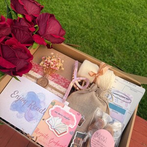 May include: A gift box filled with reading-themed items. Includes a bouquet of red roses, a bookmark, a book review sheet, a small bag, and a package of sea salt caramels. The box is on a wooden surface with a green grass background.