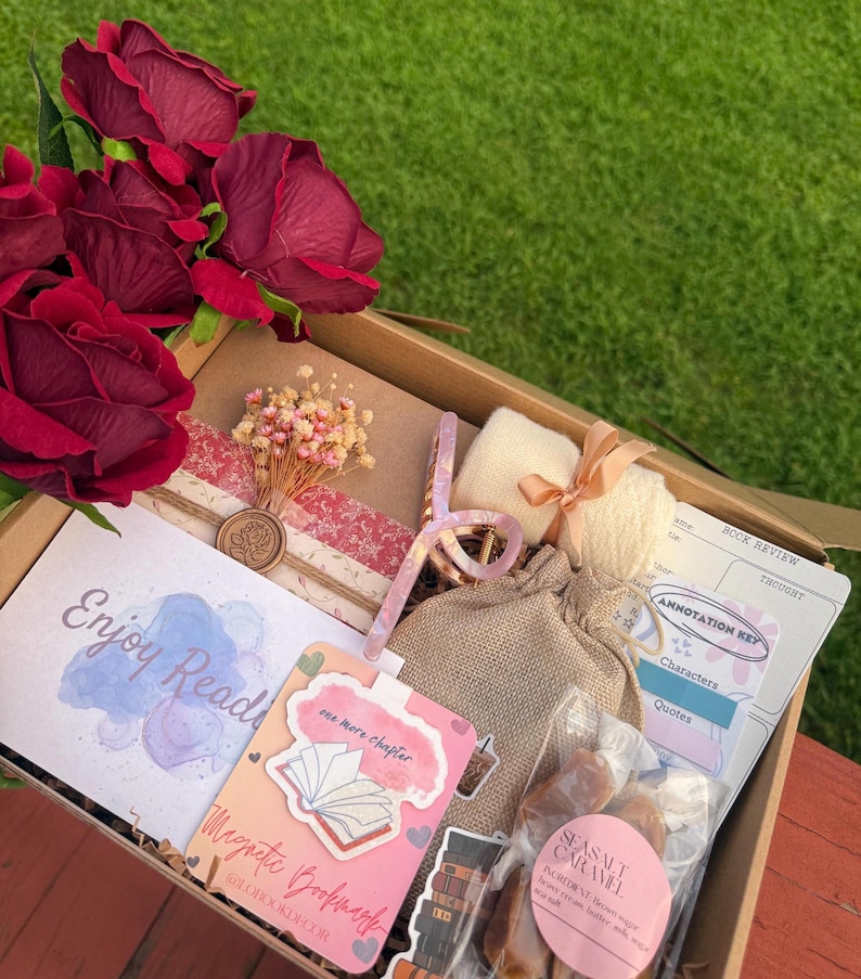 May include: A gift box filled with reading-themed items. Includes a bouquet of red roses, a bookmark, a book review sheet, a small bag, and a package of sea salt caramels. The box is on a wooden surface with a green grass background.