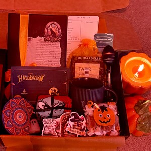 May include: A Halloween-themed gift box filled with a variety of items. Includes a book, a mug, a candle, stickers, candy, and a decorative ornament. The box is open, revealing the contents.