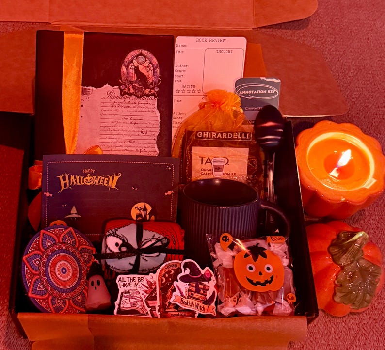 May include: A Halloween-themed gift box filled with a variety of items. Includes a book, a mug, a candle, stickers, candy, and a decorative ornament. The box is open, revealing the contents.
