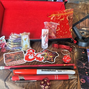 May include: A collection of Dungeons & Dragons themed items, including stickers, dice, pens, and a red tray. The stickers feature dragons and the D&D logo. The pens are red and black. A red dice keychain and a display case are also present.