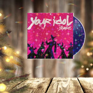 May include: A decorative ornament featuring a pink background with the text "Your Idol" and "SKINBOYS". The ornament has a silhouette of figures in black and a blue disc with star-shaped designs.