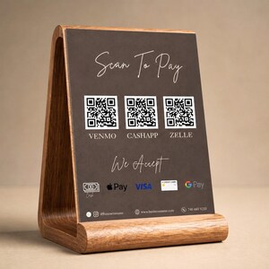A warm lifestyle product shot showing the dark brown luxury price list template displayed upright in a wooden tabletop stand with a large scan to pay QR code panel and We Accept text visible representing how the luxury price list looks as a physical desk display giving hair salon owner, wig seller, and beauty professional a premium in person payment and service menu solution