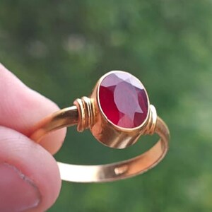 Könnte beinhalten: Ein goldener Ring mit einem großen, oval geschliffenen Rubin. Der Rubin hat eine tiefrote Farbe und ist in eine goldene Fassung gefasst. Der Ring wird vor einem verschwommenen grünen Hintergrund gehalten.
