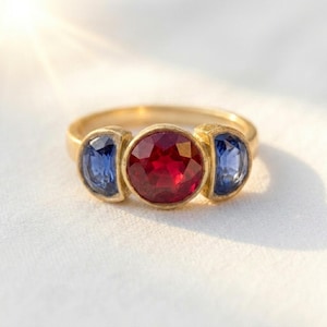 Peut inclure: Une bague en or avec une pierre précieuse rouge ronde à facettes au centre, encadrée par deux pierres bleues plus petites en forme de croissant. L'anneau est lisse et métallique, maintenant les trois pierres dans un serti clos.