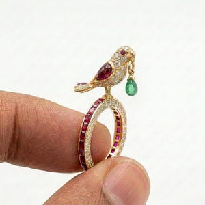 May include: A gold ring featuring a bird motif adorned with pavé diamonds and a marquise-cut ruby wing. The bird holds a pear-shaped green emerald drop in its beak. The ring band is encrusted with alternating channel-set rubies and diamonds.