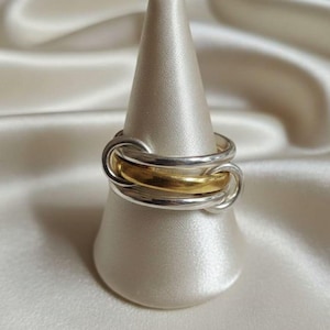 May include: A multi-band ring featuring three interlocking bands, with two polished silver-toned bands flanking a single central gold-toned band. The rings are connected in a continuous loop design, creating a layered appearance on a conical display.
