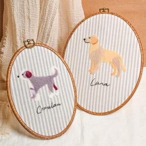 Op de afbeelding: Twee ovale borduurringen met hondenportretten. Eén toont een paarse en witte hond, de andere een golden retriever. Beide staan op gestreepte stof, met de namen van de honden eronder geborduurd.