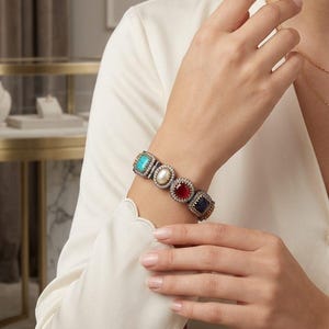 May include: A silver bracelet featuring five square gemstones: turquoise, pearl, ruby, and sapphire. The bracelet is worn on a wrist with a white long-sleeved shirt.