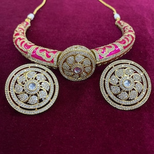 May include: A gold-toned jewelry set featuring a necklace and matching earrings. The necklace has a pink and gold design with a central circular pendant. The earrings are circular with a floral pattern and clear stones.