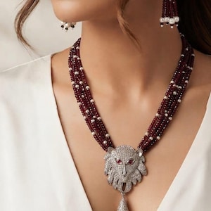 May include: A multi-strand necklace featuring deep red beads and small white pearls, centered with a silver-toned lion head pendant encrusted with clear stones and red eyes, finished with a matching tassel and coordinating drop earrings.