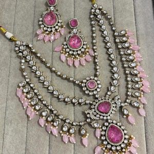 May include: A three-piece jewelry set featuring a necklace and matching earrings. The set is silver-toned with clear and pink gemstones. The necklace has multiple strands and a large pink center stone. The earrings have a teardrop shape.