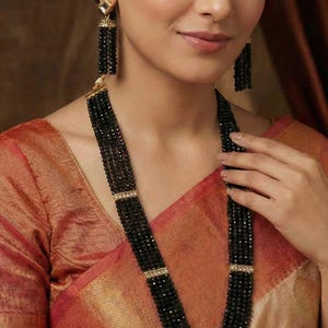 May include: A black beaded necklace and matching earrings set. The necklace features multiple strands of small, dark beads with gold-colored accents. The earrings have a similar design, with a square top and dangling beads. The jewelry is worn with a red and gold sari.