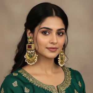 May include: Gold-toned statement earrings with a rectangular top, a maroon center, and a crescent-shaped bottom adorned with pearls. The earrings feature green and gold accents. The woman is wearing a green top with gold embroidery.