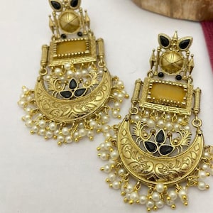 Puede incluir: Un par de pendientes dorados con detalles intrincados. Cada pendiente presenta una piedra amarilla rectangular, detalles en negro y un diseño en forma de media luna adornado con pequeñas perlas blancas. Los pendientes tienen una estética vintage.