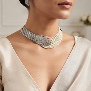 Puede incluir: Un collar tipo gargantilla de múltiples hilos con una combinación de perlas blancas y cristales transparentes, con un diseño asimétrico que transita de filas incrustadas de cristales a hilos de perlas, junto a pendientes de botón a juego.
