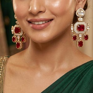May include: Gold-toned chandelier earrings with large, rectangular, ruby-red gemstones. The earrings feature smaller, rectangular red stones and clear, round stones. The earrings are worn with a dark green garment.