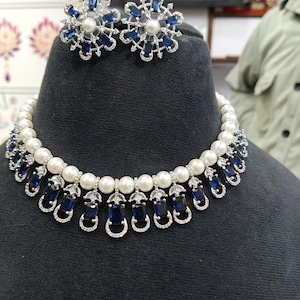 Puede incluir: Un conjunto de collar y pendientes exhibido en un maniquí negro. El collar presenta perlas blancas y piedras azules rectangulares. Los pendientes tienen forma de copo de nieve con piedras azules y una perla central. La joyería es de plata.
