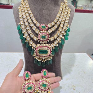 Op de afbeelding: Een goudkleurige ketting en bijpassende oorbellen set. De ketting heeft meerdere lagen witte edelstenen en smaragdgroene druppelvormige hangers. De oorbellen en ketting hebben smaragdgroene en robijnrode accenten.