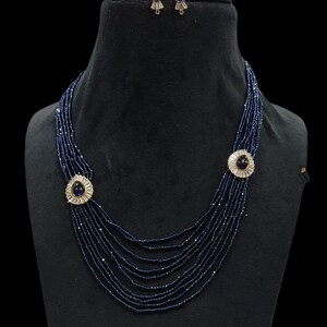 Può includere: Un set di collana e orecchini con perline blu navy. La collana presenta più fili di piccole perline, con due chiusure circolari e ornate. Ogni chiusura ha una pietra centrale blu scuro circondata da pietre più piccole e trasparenti. Gli orecchini hanno un design simile.