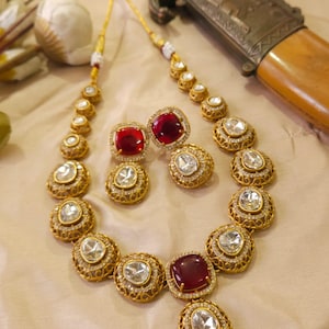 May include: A gold-toned necklace and matching earrings set with clear and red gemstones. The necklace features a series of round and oval-shaped stones, while the earrings have square-shaped red stones. The jewelry is displayed on a light-colored surface.