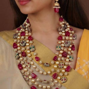 May include: A multi-strand necklace and matching earrings set. The necklace features layers of pearls, red gemstones, and ornate gold-toned metalwork. The earrings have a similar design with red gemstones and gold accents. The jewelry is displayed on a person.