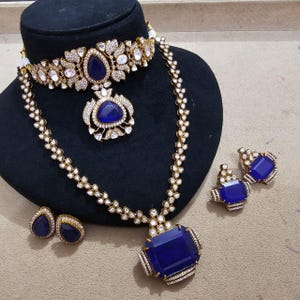 May include: A gold-toned jewelry set featuring a choker necklace with a central teardrop blue stone, a long necklace with a large rectangular blue pendant, and two pairs of matching earrings, all accented with small white stones in a floral pattern.