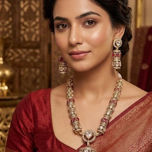 May include: A gold-toned necklace and matching drop earrings featuring embedded red oval stones and clear white crystals. The necklace includes a large cylindrical pendant with a central teardrop-shaped stone, all set in intricate metalwork.