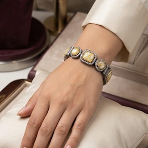 May include: A silver and gold bracelet with square and round yellow stone accents. The bracelet is set with small, clear stones around the yellow stones. The bracelet is on a person's wrist, resting on a white pillow.