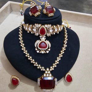 Puede incluir: Un conjunto de joyas de tono dorado que incluye un collar, pendientes y un anillo. El collar tiene un colgante grande, rectangular, de piedra roja y un gargantilla a juego. Los pendientes y el anillo también presentan piedras rojas. El conjunto está adornado con pequeñas piedras preciosas transparentes.