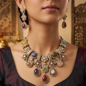 May include: A gold-toned statement necklace and matching drop earrings featuring an array of multi-colored cabochon gemstones including purple, red, green, blue, and yellow stones, accented with small clear crystals set in intricate metalwork.