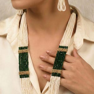 May include: An elegant necklace and earring set. The necklace features strands of white beads with dark green and gold accents. The earrings have a gold top with white beaded tassels. The jewelry is worn over a cream-colored blouse.