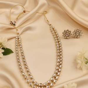 May include: A gold-coloured necklace and matching earrings set. The necklace has two strands of oval, clear stones. The earrings are flower-shaped with clear stones. The jewellery is displayed on a cream-coloured, satin-like fabric.