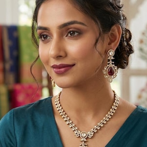 Puede incluir: Un conjunto de joyas doradas con collar y pendientes. El collar tiene un colgante central con una gran piedra preciosa roja, rodeada de piedras más pequeñas y transparentes. Los pendientes reflejan el diseño del colgante. La mujer lleva una blusa color turquesa.