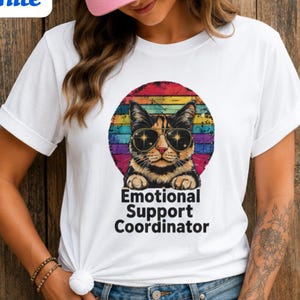 May include: White t-shirt with a cat wearing sunglasses, set against a rainbow-striped background. The text "Emotional Support Coordinator" is below the cat image. The word "White" is in a blue box in the upper left corner.