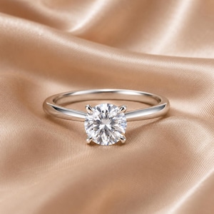 May include: A silver-toned ring featuring a single round, brilliant-cut clear stone set in a four-prong setting. The band is smooth and polished, showcasing a reflective metallic finish that complements the faceted center stone.