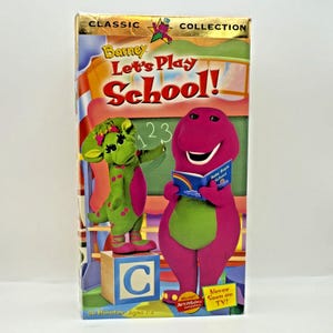 May include: A VHS tape from the Classic Collection featuring Barney the dinosaur. The cover shows Barney and Baby Bop in a classroom setting with the text "Let's Play School!" and "Never Seen on TV!" The tape is labeled for ages 1-3.