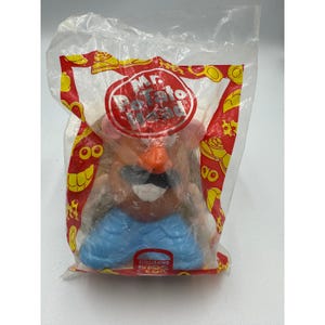 May include: A Mr. Potato Head toy figure sealed in a clear plastic bag with red and yellow graphics. The figure has a brown body, orange nose, black mustache, white teeth, and blue pants. The packaging features the Mr. Potato Head and Burger King Kids Club logos.