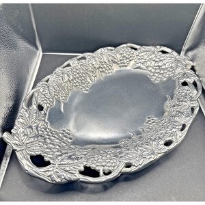 May include: Oval silver-toned serving dish with a grape and leaf design around the rim. The dish has a smooth, reflective surface and is likely made of metal. The grape and leaf pattern adds a decorative touch to the serving piece.