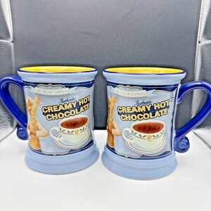 May include: Two ceramic mugs with blue handles and bases, featuring the text "Authentic Creamy Hot Chocolate" and a graphic of a cup of hot chocolate. The mugs have a yellow rim and a light blue body. The Polar Express is written on the top.