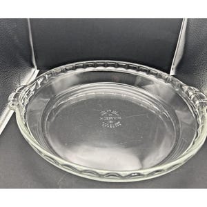 May include: Clear glass pie dish with a fluted edge and two handles. The dish has a circular shape and is made of transparent glass. The Pyrex logo is visible in the center of the dish.