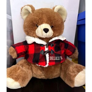 May include: A brown plush teddy bear wearing a red and black buffalo plaid jacket with a white sherpa collar. The jacket features dark toggle closures and the embroidered text BELKIE '94 on the front. The bear has cream-colored paw pads.